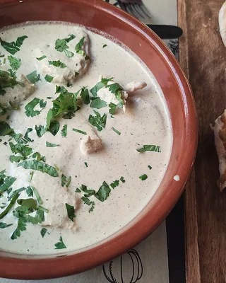 Chicken satsivi in a clay bowl under a walnut sauce with herbs, a loaf of bread sliced beside it