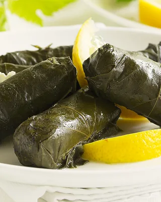 Vine leaf tolma on a white plate with lemon wedges and a jug of yoghurt sauce