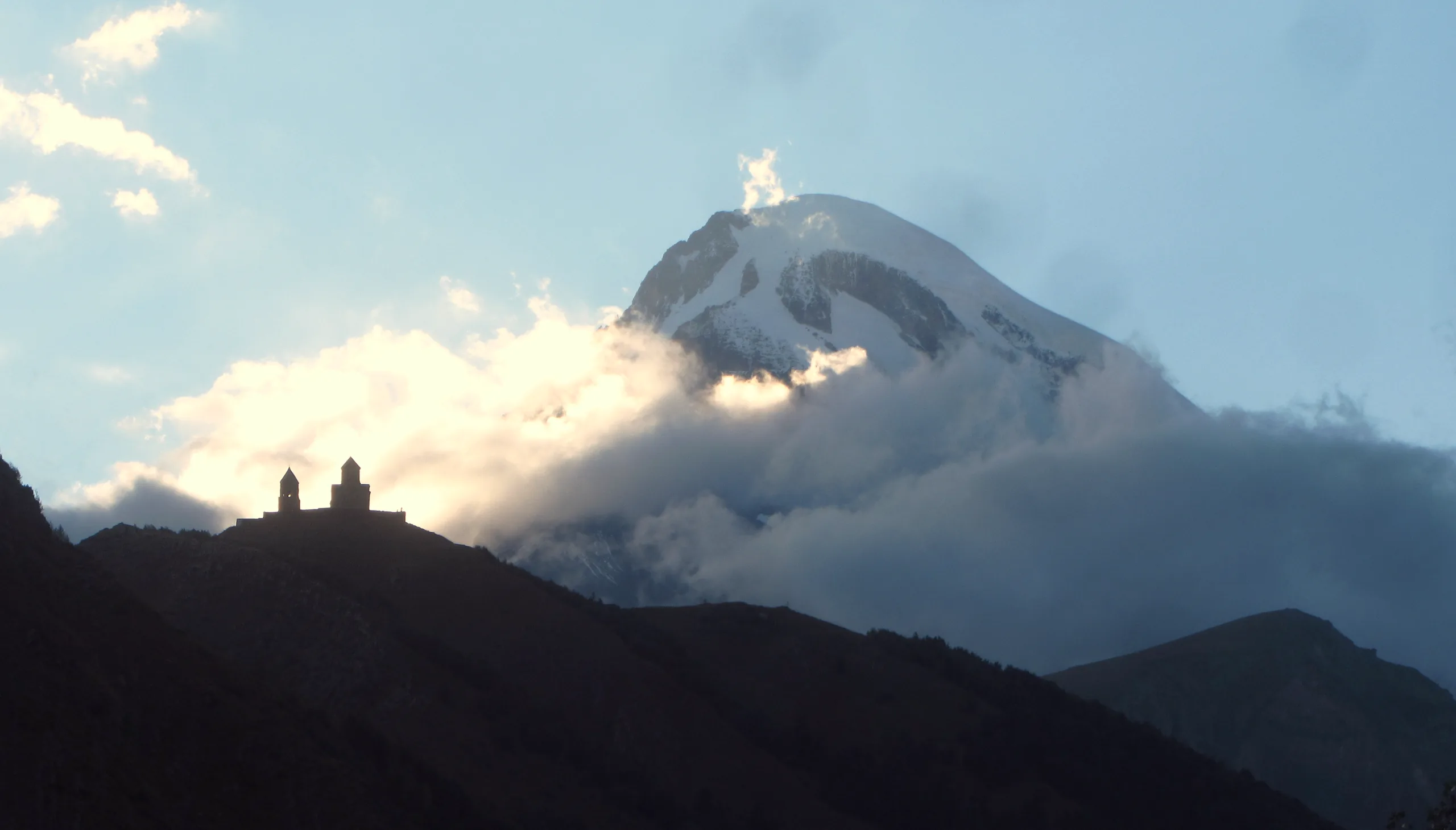 Gergeti Trinity Church in silhouette on its ridge, cloud tearing off the snow face of Mount Kazbek behind it at dusk