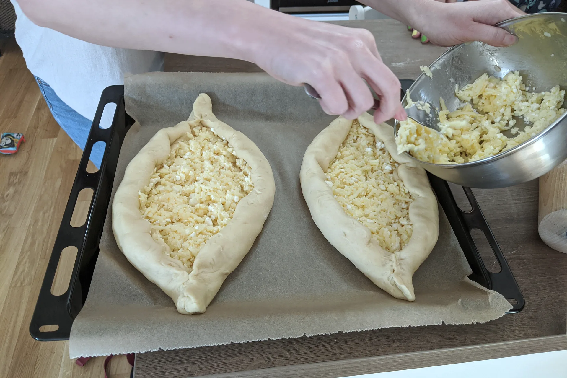 Hands spooning cheese into two Adjaruli khachapuri boats on a baking tray