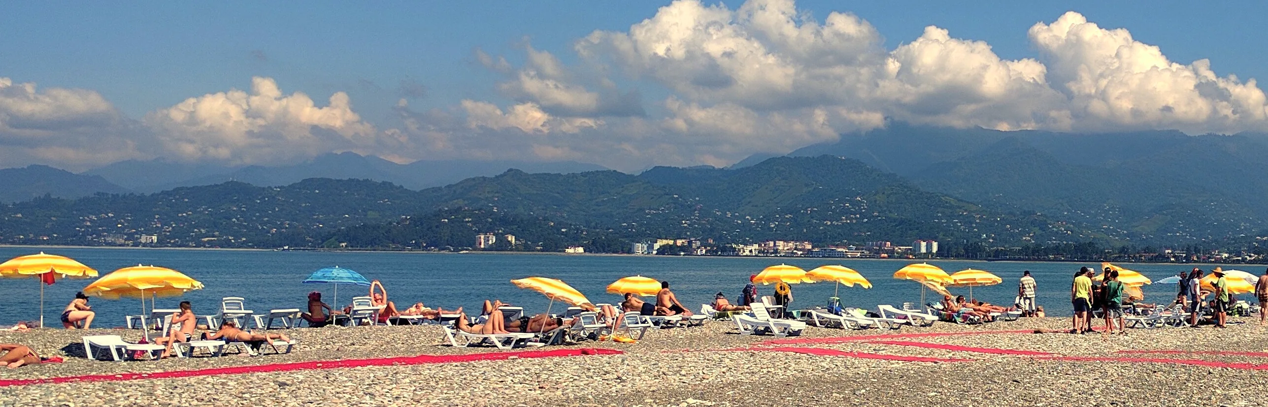 Yellow umbrellas and sunbathers on the beach at Batumi, blue sea and green hills behind, in full sun