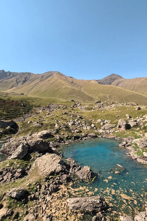 A small turquoise lake among grey rocks and green slopes under a blue sky