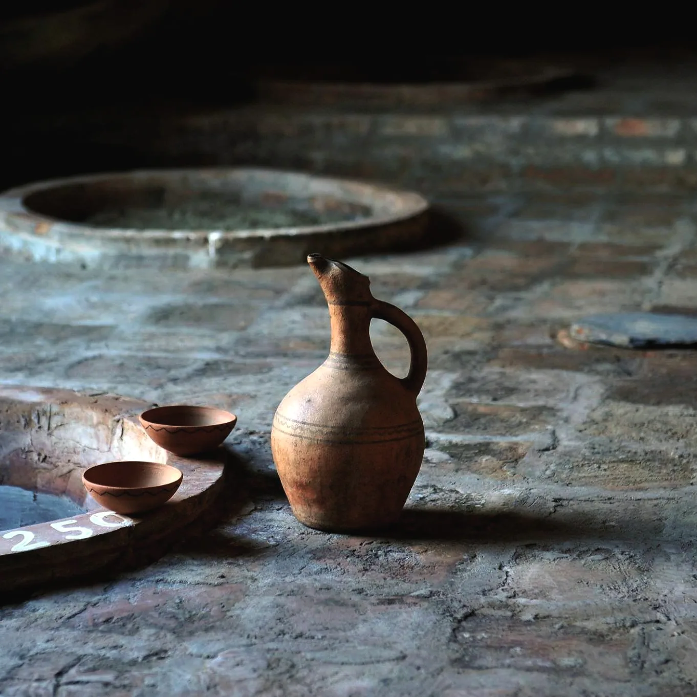 A clay wine jug and two clay bowls on the rim of a sunk qvevri in a stone marani, in raking light